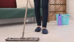 Vrouw dweilt vloer in woonkamer met emmer en schoonmaakmiddel op de achtergrond.