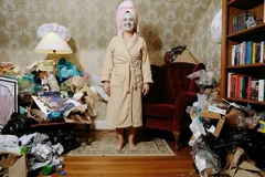 Vrouw in badjas tussen rommel in een overvolle, slordige woonkamer met boekenplank.