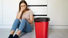 Vrouw zit verdrietig naast rode prullenbak in keuken, hand voor mond, emotioneel moment.