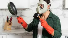 Man met gasmasker en oranje handschoenen houdt pan vast in keuken