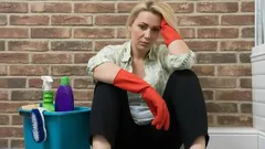 Vermoeide vrouw met schoonmaakmiddelen en rode handschoenen zittend op de vloer tegen een muur.