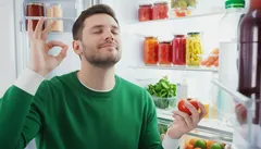 Man geniet van gezonde voeding bij open koelkast, houdt tomaat vast, maakt tevreden gebaar.