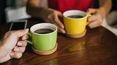 Twee mensen drinken koffie uit gekleurde mokken aan houten tafel in café.