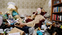 Vrouw in badjas zit in rommelige kamer vol afval en boeken, leest en drinkt koffie.